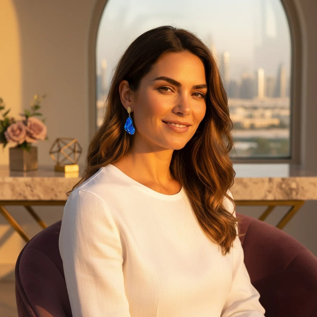 Model wearing The Accessory Bar gold-plated geometric drop earrings with bold blue cubic zirconia in a sunset setting.